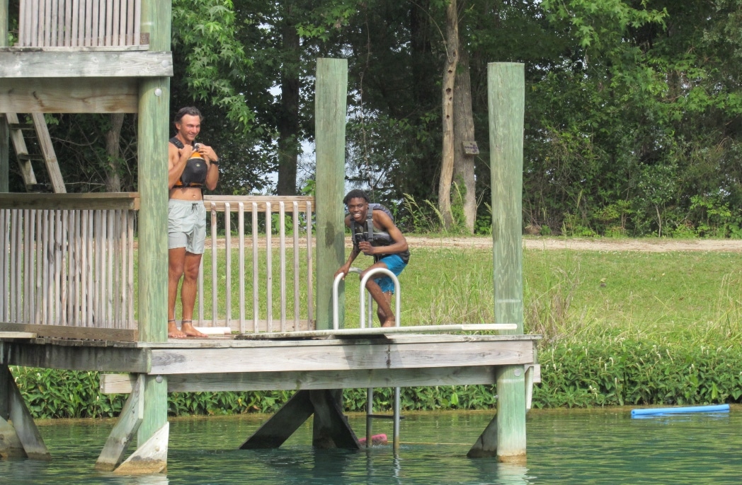 Camp Photo 2 edit Two young people stand on a dock near a body of water with life jackets on during a summer camp.
