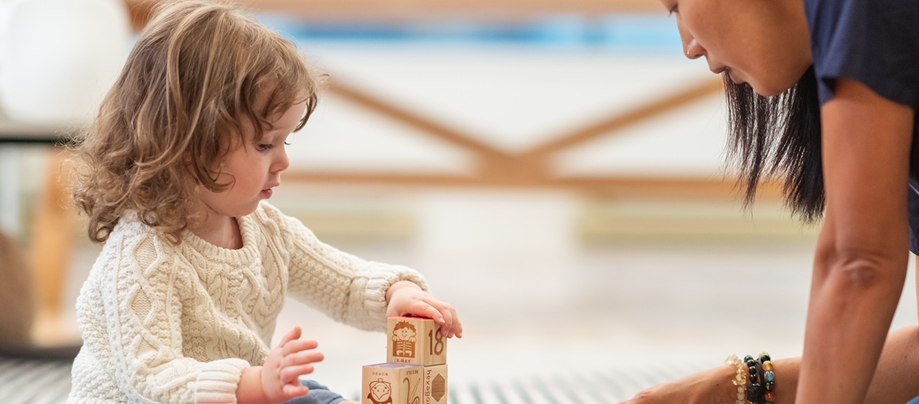 A preschool age girl with a prosthetic leg is at a medical appointment. The child is meeting with her physical therapist. The child is sitting on the floor building with wooden toy blocks. The medical professional is sitting on the floor assisting the girl.