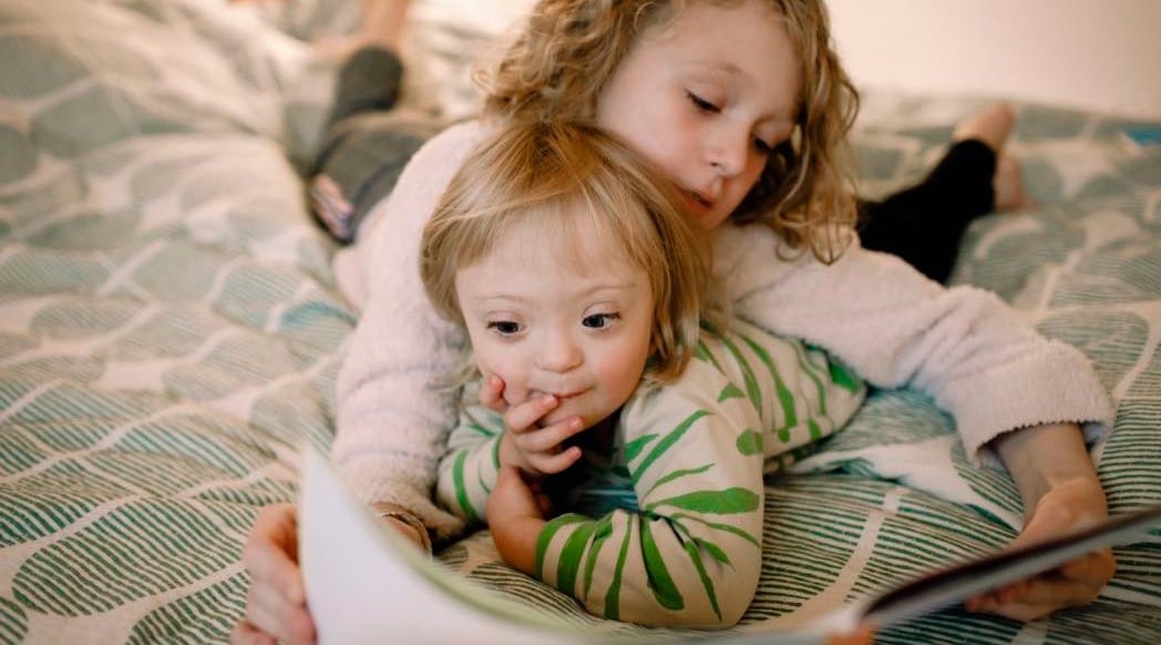 Female sibling reading book while lying with disabled sister on bed