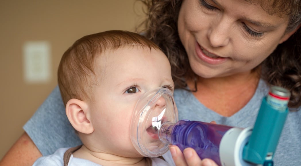 A child with asthma uses an inhaler while held by their mother.