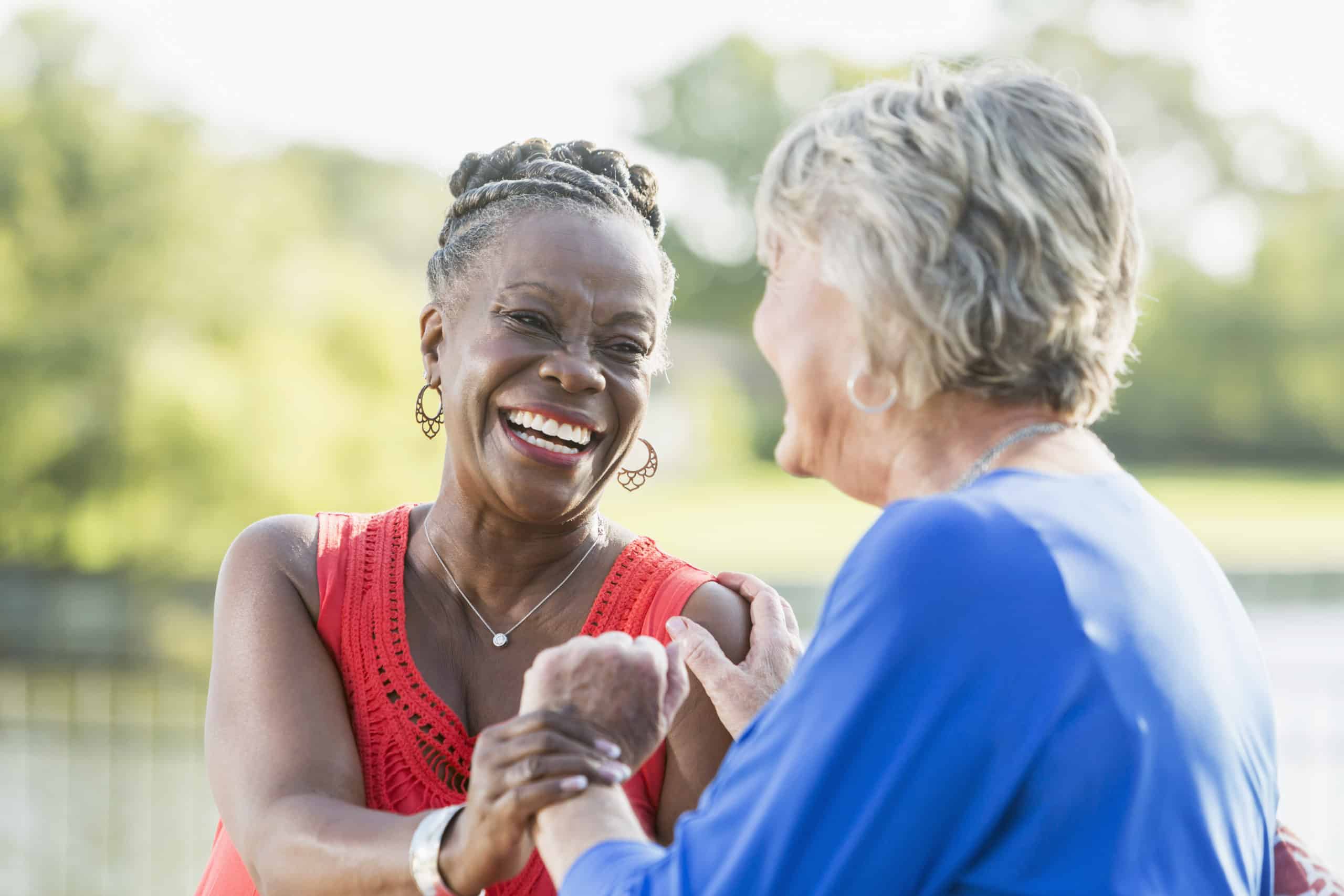 Two senior multi-ethnic woman, one Caucasian and the other African American, sitting in back yard by water enjoying each other's company. They are looking at each other, laughing.