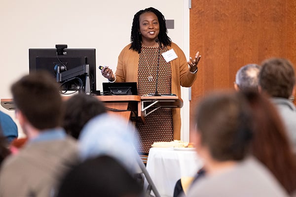 Dr. Sharona Johnson gestures to a crowd during a presentation on narrative medicine during the Albernaz Golden Apple Distinguished Lecture at East Carolina Heart Institute. Dr. Johnson is wearing a brown sweater over a patterned dress.
