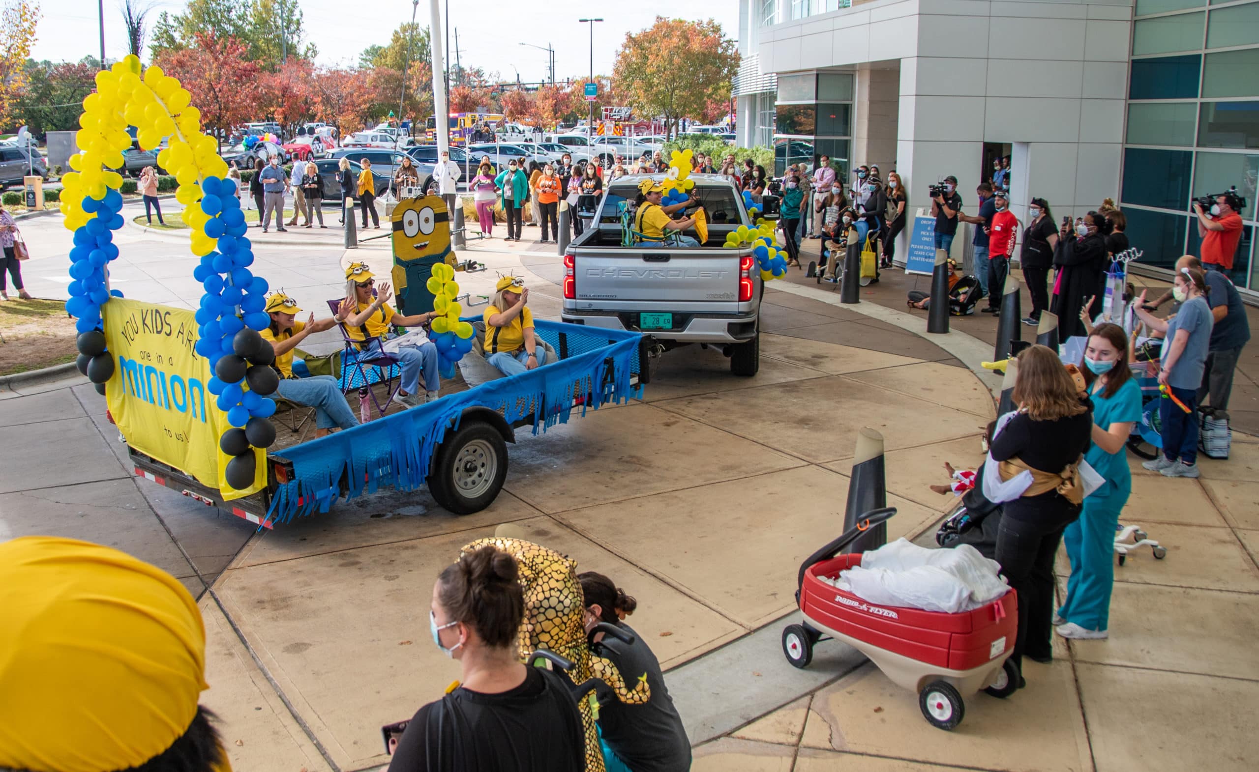 ChildrensHalloweenParade-2022-3757 A truck pulls a trailer of ECU Health team members dressed as Minions during the Halloween Parade at Maynard Children's Hospital. The truck and trailer drives through the loop in front of the Maynard Children's Hospital. Along the outside, patients, families, team members and community members wave to the team members on the float.