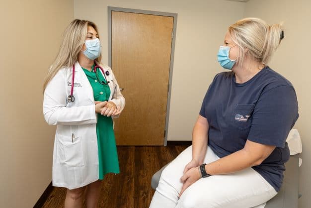 An ECU Physician speaks to a patient sitting on an examination table. They are both wearing masks and smiling while they talk.