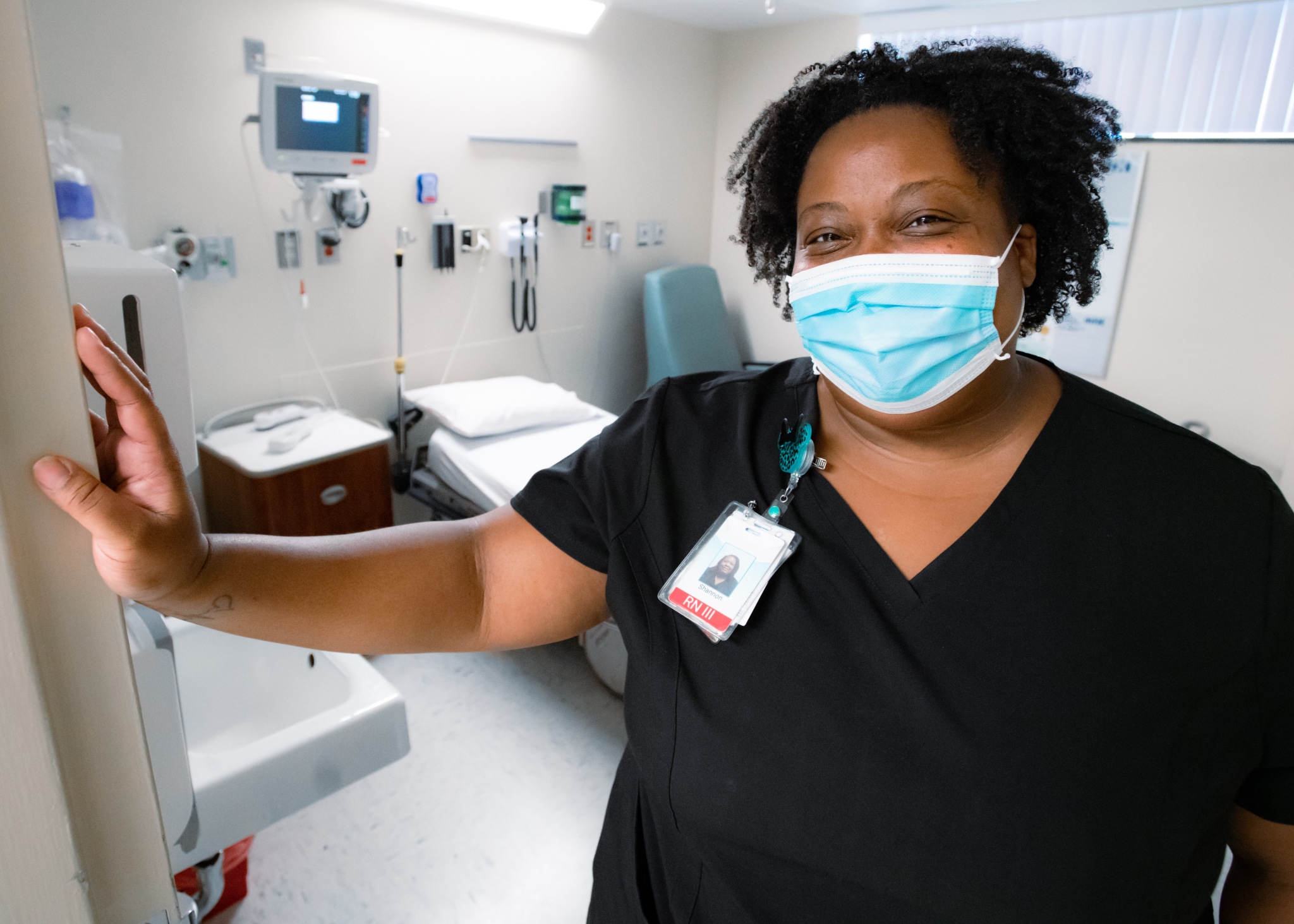 Nurse Shannon Moore poses for a photo in the doorway of an empty patient room, smiling with a mask on.