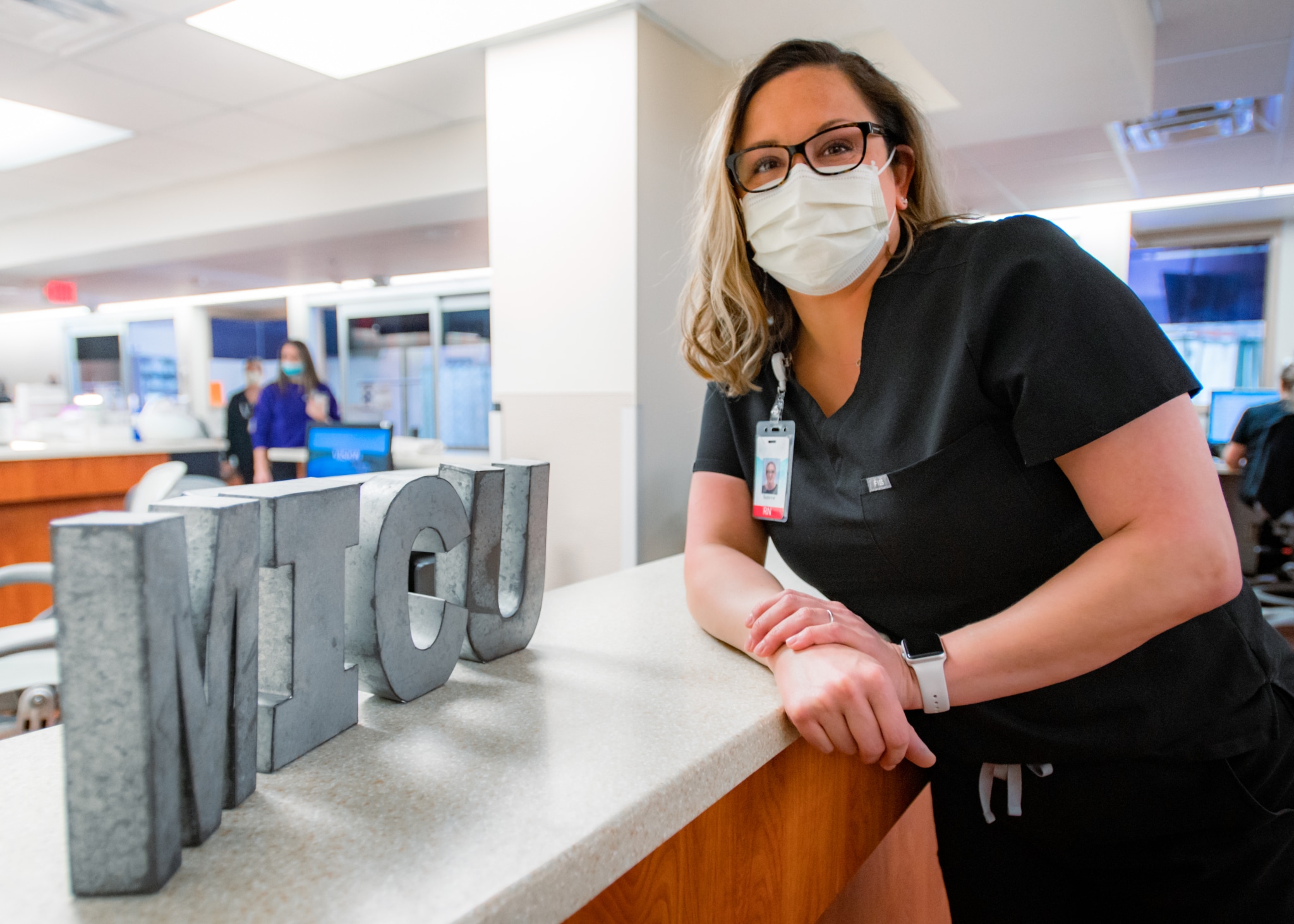 Suzanne Foster-3048 Nurse Suzanne Foster leans against a nurses station in the MICU, smiling with a mask on.