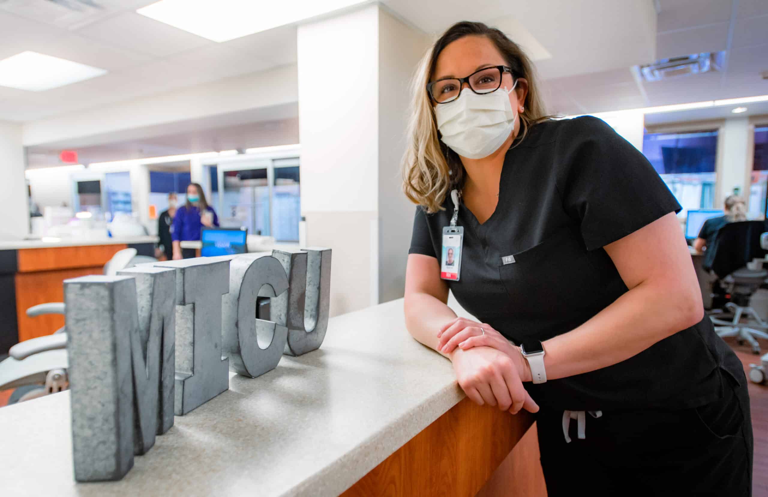 Nurse Suzanne Foster leans against a nurses station in the MICU, smiling with a mask on.