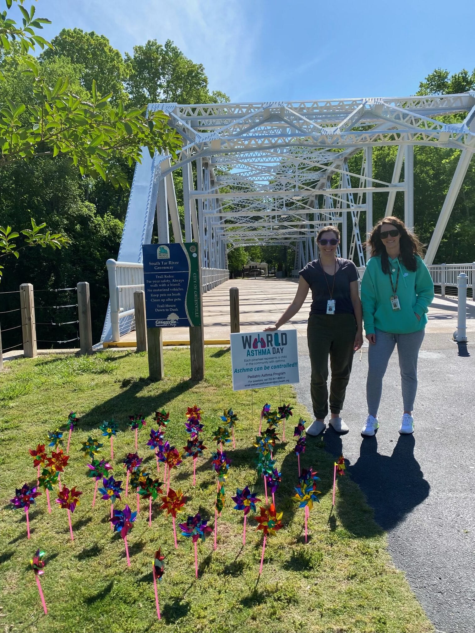Grass leading to a walking bridge in Greenville is decorated with small pinwheels, each one recognizing a pediatric asthma patient seen at Maynard Children's Hospital. Two ECU Health team members stand on the walking path, smiling for the photo.