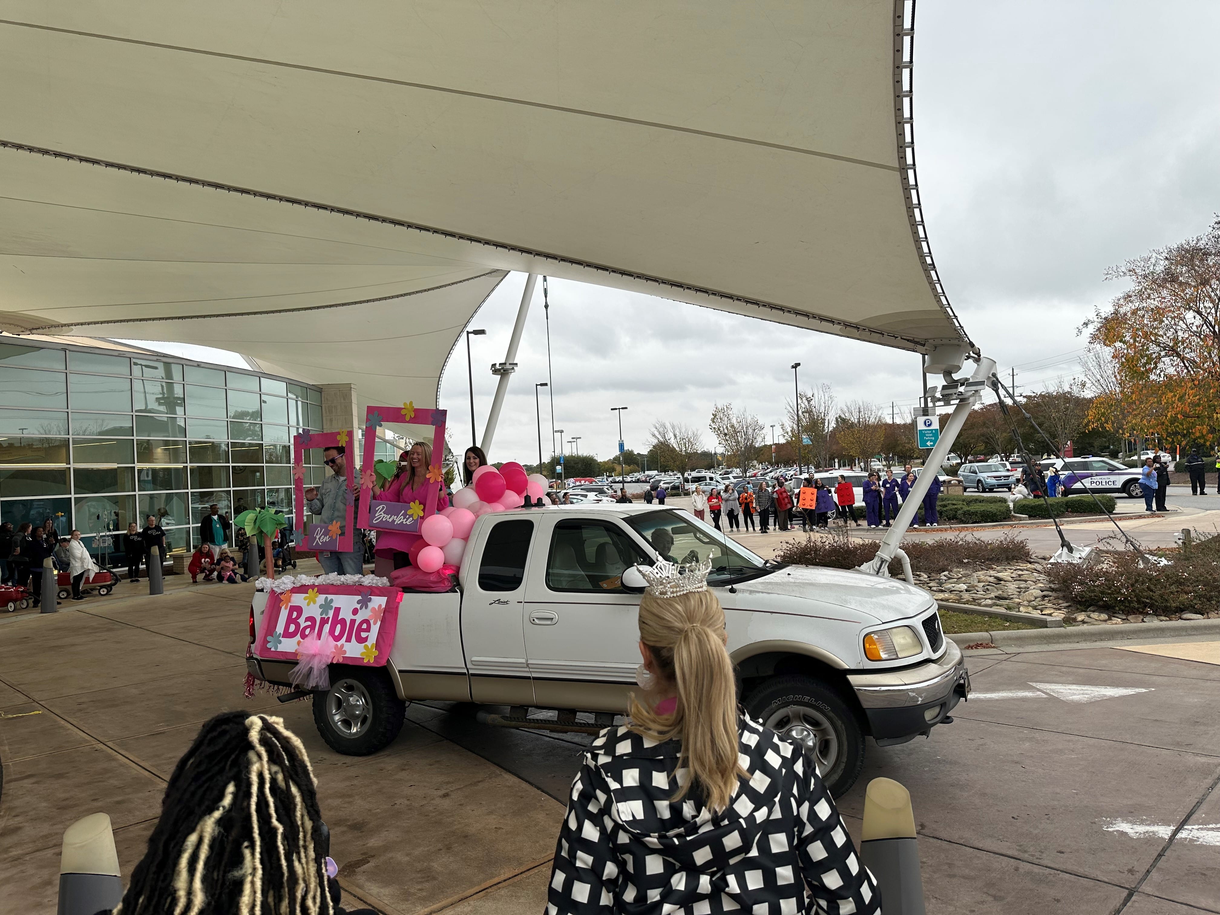 Excited ECU Health team members, dressed as Barbie and Ken, wave to Children's Hospital Halloween parade attendees from the back of a pickup truck, decked out in pink Barbie flare.