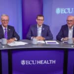 ECU Health Chief Financial Officer Andy Zukowski sits between CEO Dr. Michael Waldrum, left, and COO Brian Floyd, right, during a Looking Forward with Leadership session with ECU Health team members. The three sit behind a branded ECU Health desk.