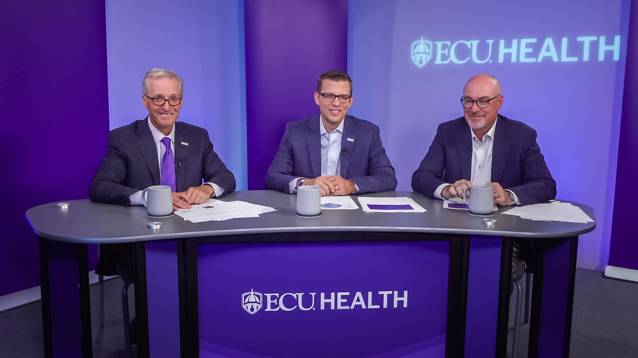ECU Health Chief Financial Officer Andy Zukowski sits between CEO Dr. Michael Waldrum, left, and COO Brian Floyd, right, during a Looking Forward with Leadership session with ECU Health team members. The three sit behind a branded ECU Health desk.