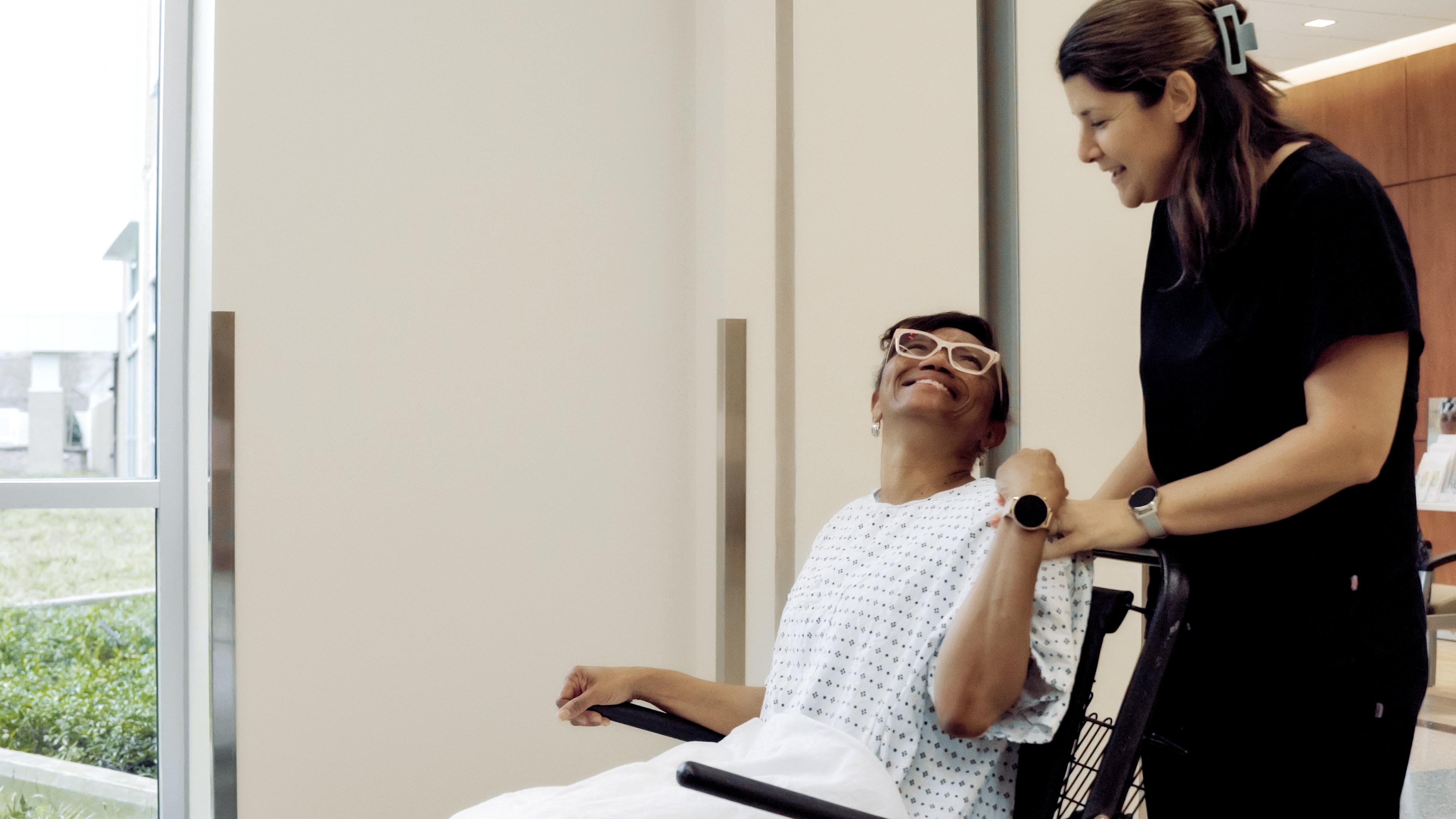 An ECU Health team member moves a patient to a room. The patient sits in a wheelchair and smiles up at the care team member.