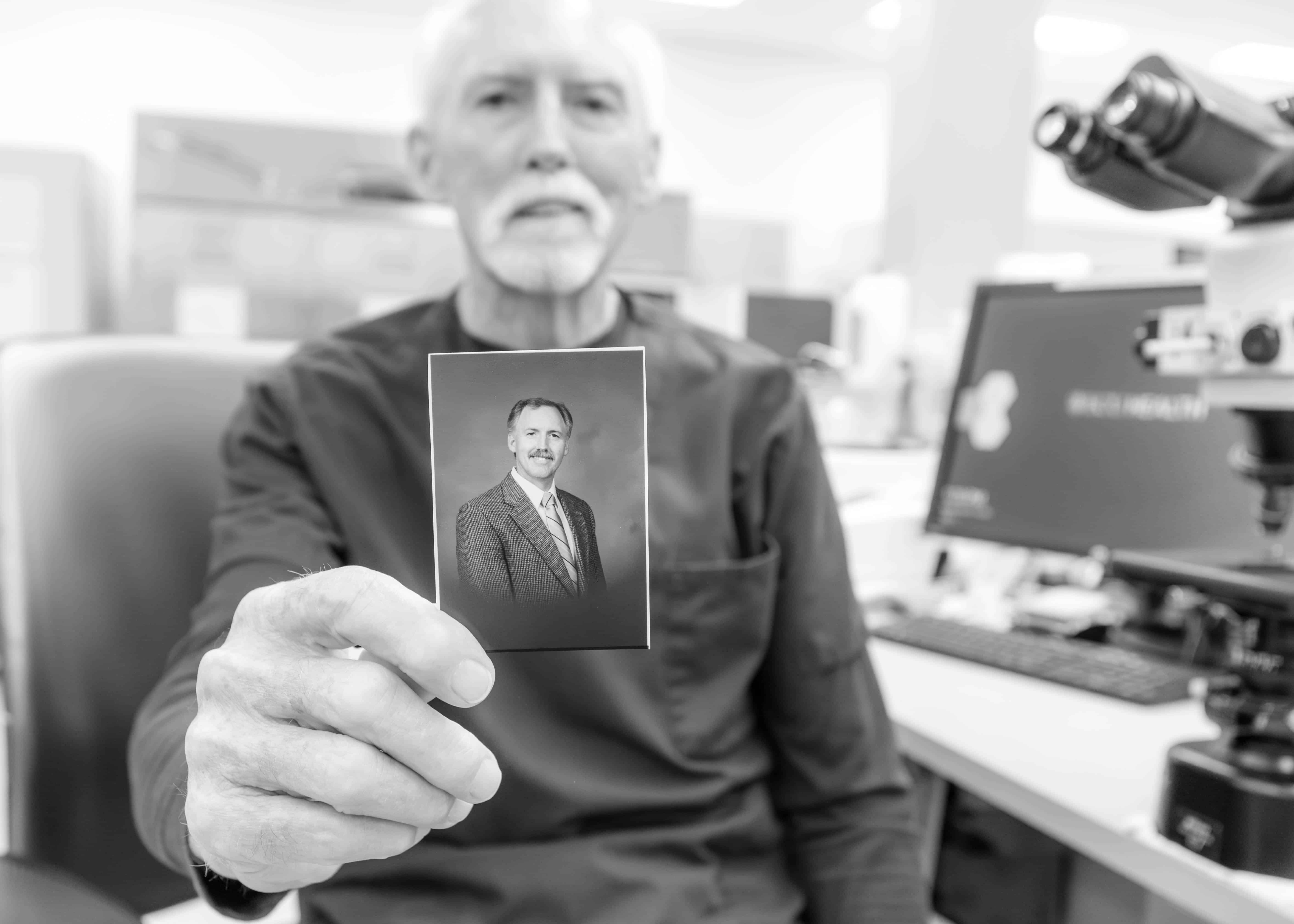 Berry Warren sits at his desk and holds a picture of himself during his early days at ECU Health.