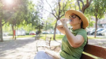 A woman sits in shade and drinks water on a park bench during a warm summer day.