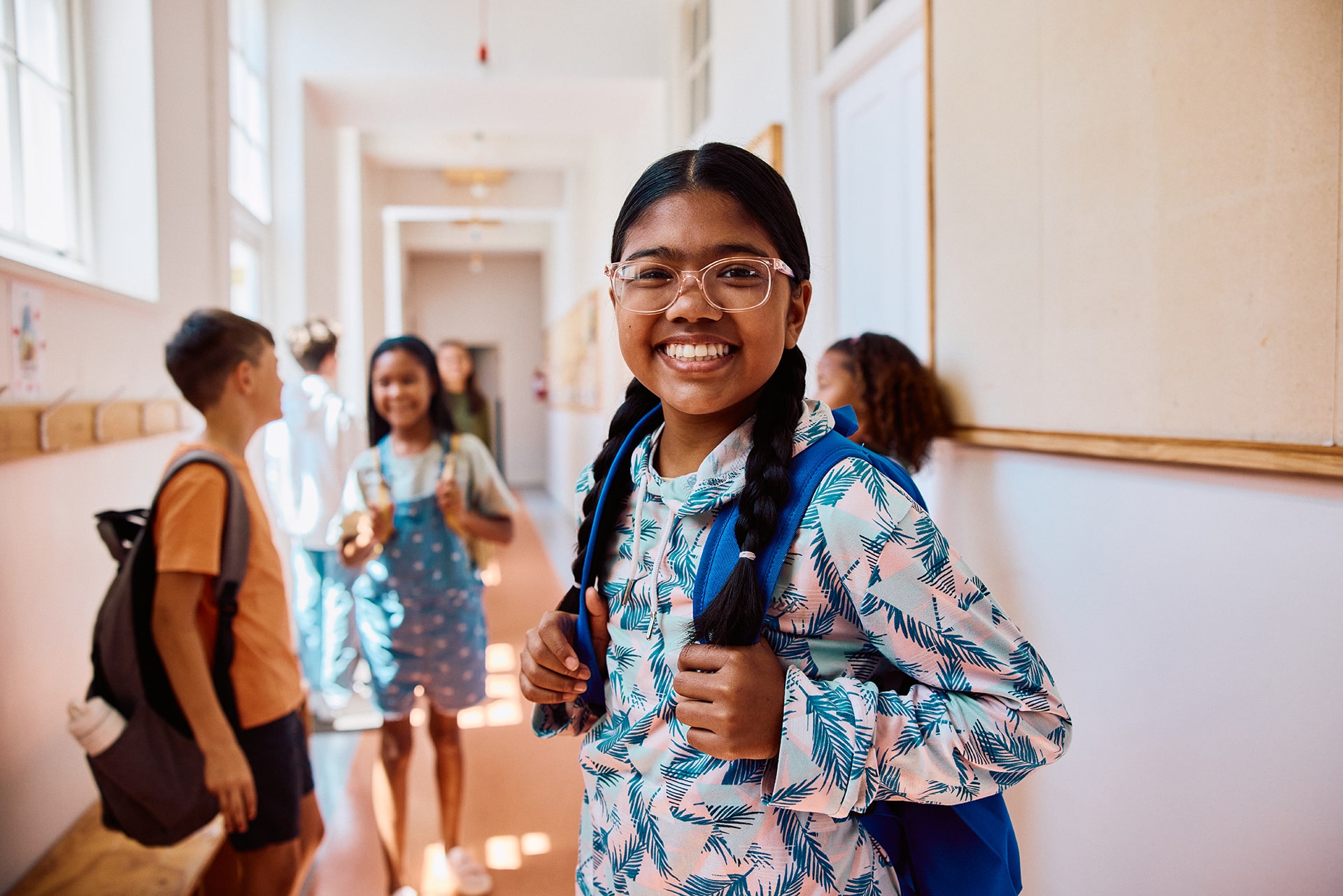 Portrait of happy school girl with glasses and braided hair smiling at camera in hallway with backpack